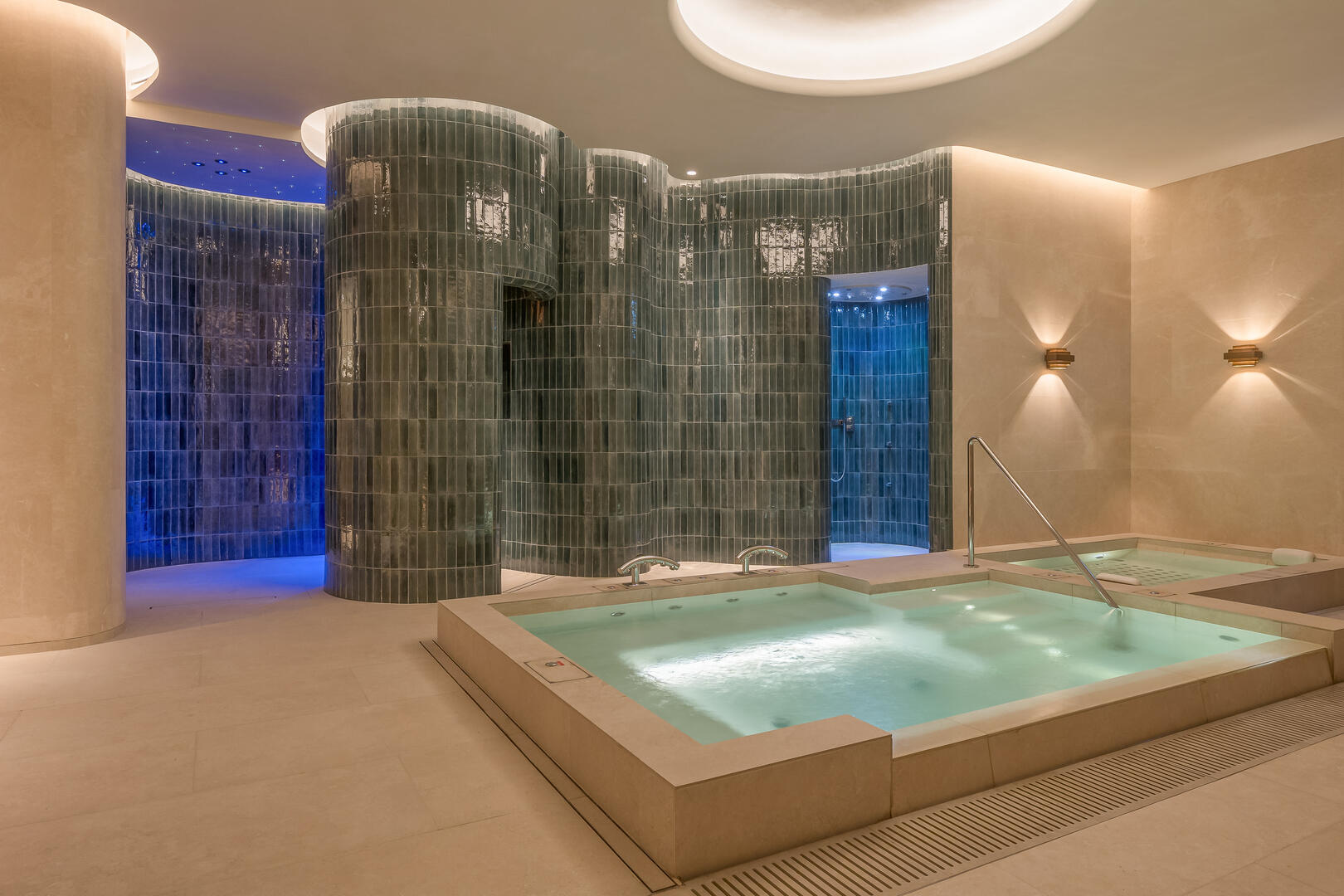 Luxurious indoor spa featuring a warm hydrotherapy pool with built-in jets and modern fixtures. The space is accentuated by curved emerald green tiled walls leading to sensory showers with ambient lighting, creating a tranquil and rejuvenating wellness environment.